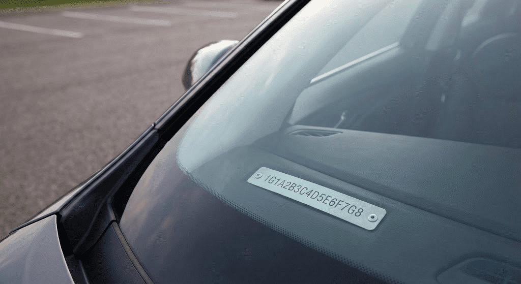 View from outside a car looking through the windshield at the 17-character Vehicle Identification Number (VIN) plate on the dashboard. Placement: Inside the "What Is VIN?" section, after the "What a VIN Check Reveals" sub-section.